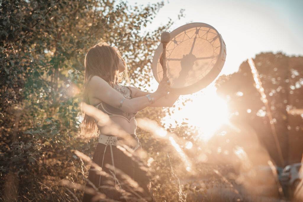A traditional slavic shaman girl beating the drum, evoking the spirits of nature in a lovely sunset field scenery as the trance dance ritual